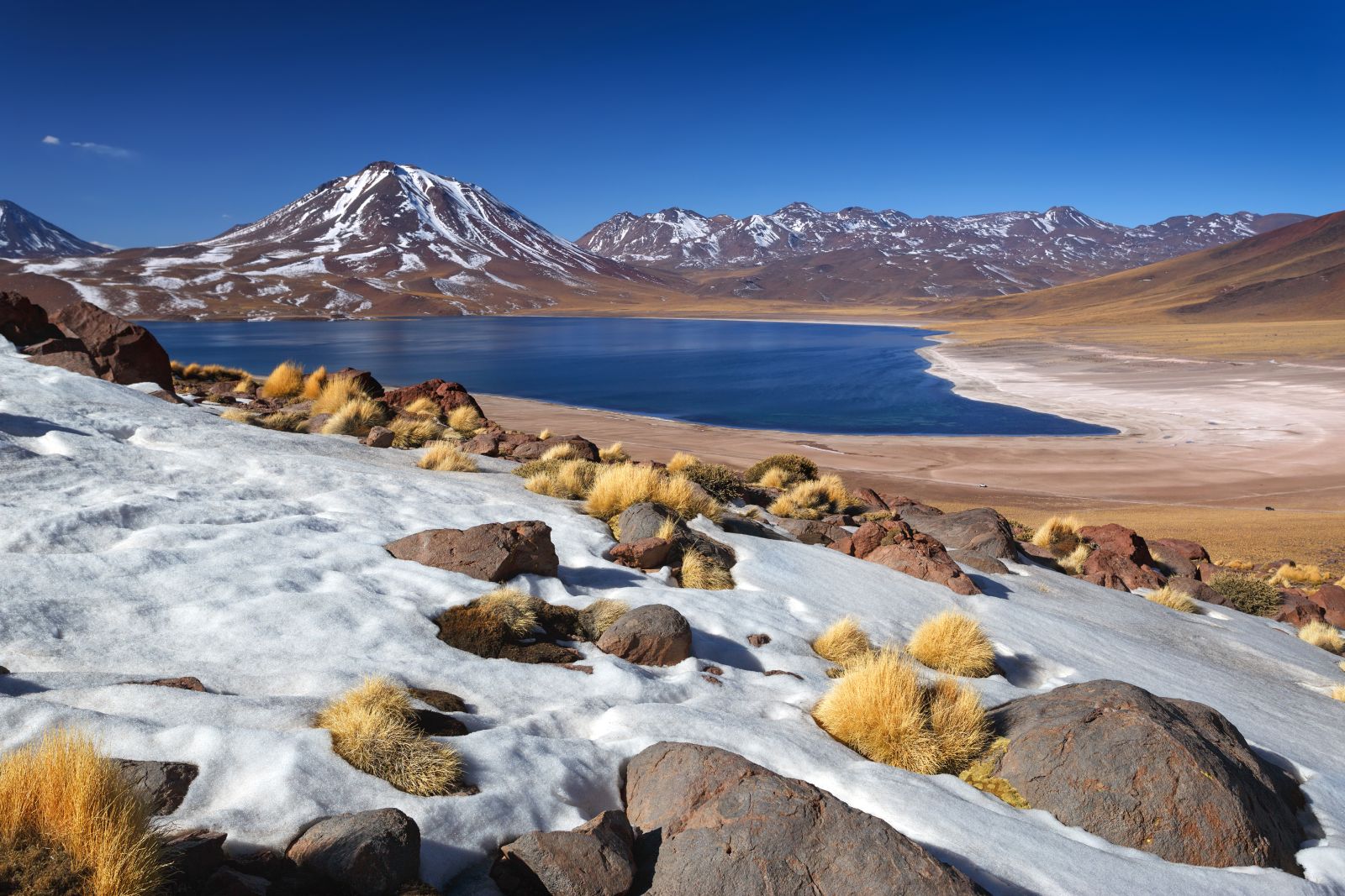 A layer of snow in the Atacama Desert in Chile with a blue lake and volcano in the background