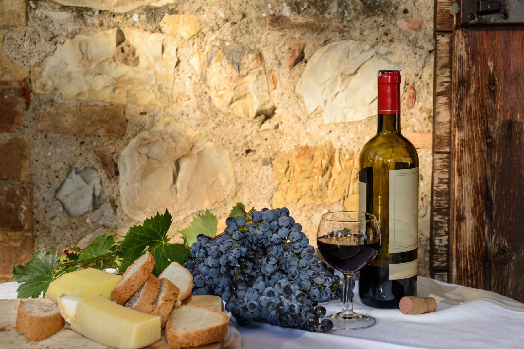 A bottle of red wine on a table with grapes, cheese and fresh bread