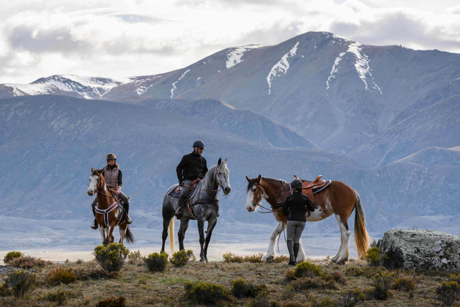 Two people sat on horses and one person holding a horse in the mountains at The Lindis Lodge in New Zealand