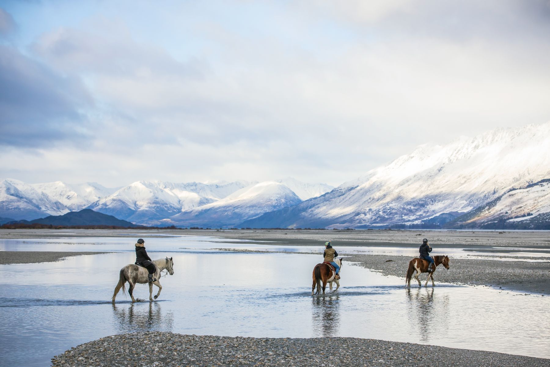 People horse riding on an empty beach in Glenorchy New Zealand with snowcapped mountains